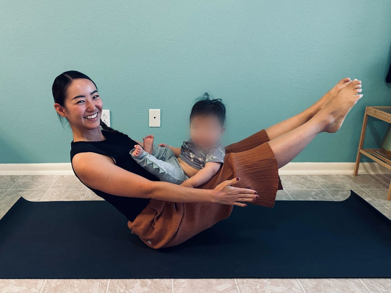 A mother smiling while balancing in boat pose with her baby held against her legs on a yoga mat at home