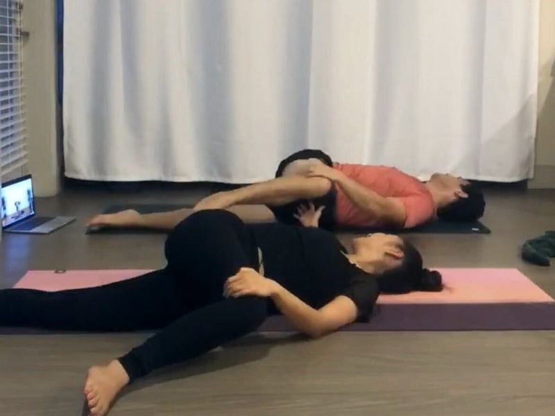 A couple practicing a supported reclined twist together during an online session, laptop visible